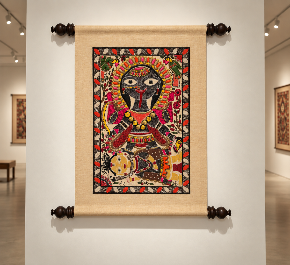 A traditionalmadhubani folk art scroll painting mounted on a gallery wall. The scroll has a beige linen border and dark wooden finials at the corners, displaying a colorful painting of a goddess kali with red and yellow details, set against a white gallery background with other art pieces visible.